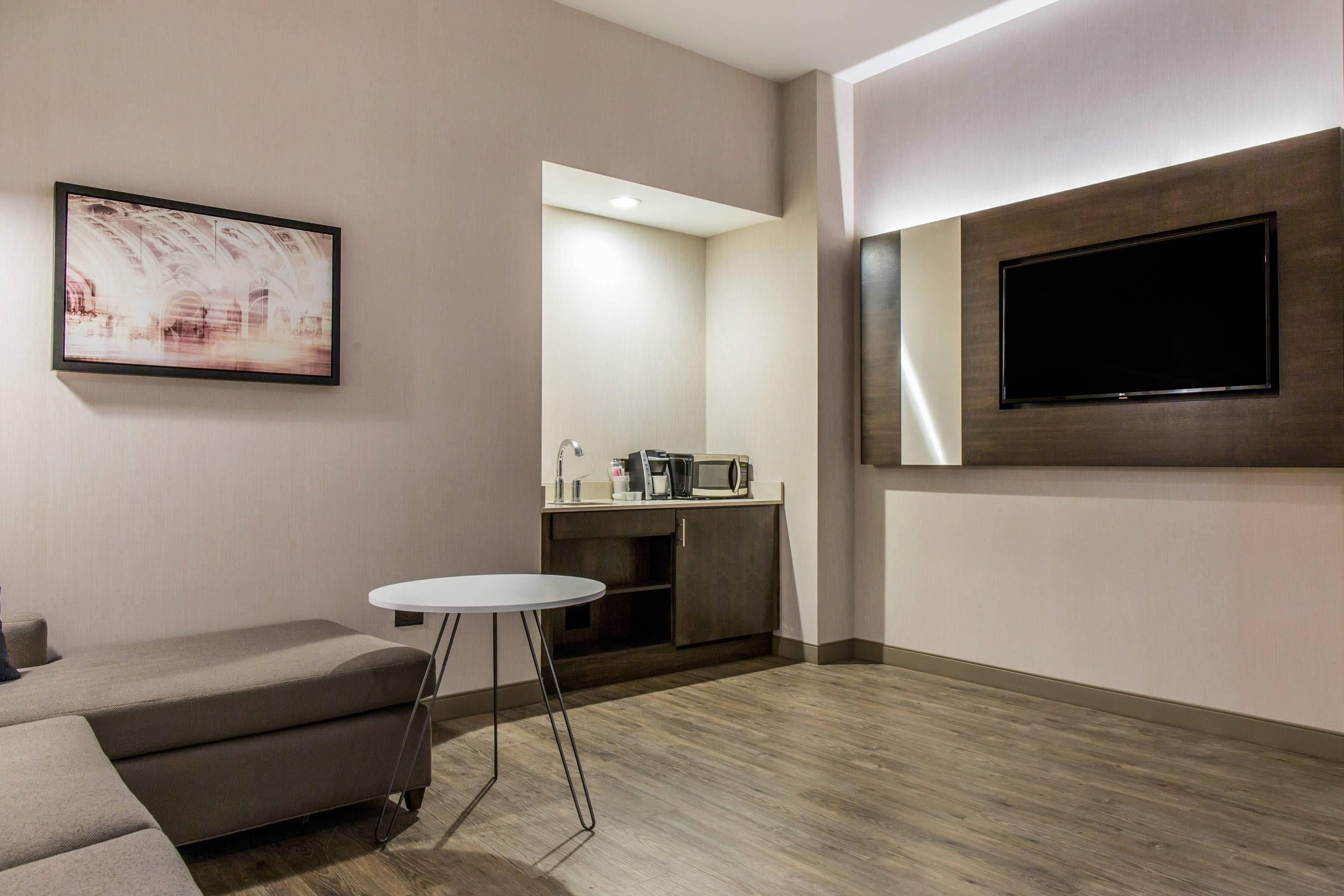 A modern living room with a grey sofa, a small round table, a flat-screen TV mounted on the wall, and a kitchenette area with a microwave and coffee maker.