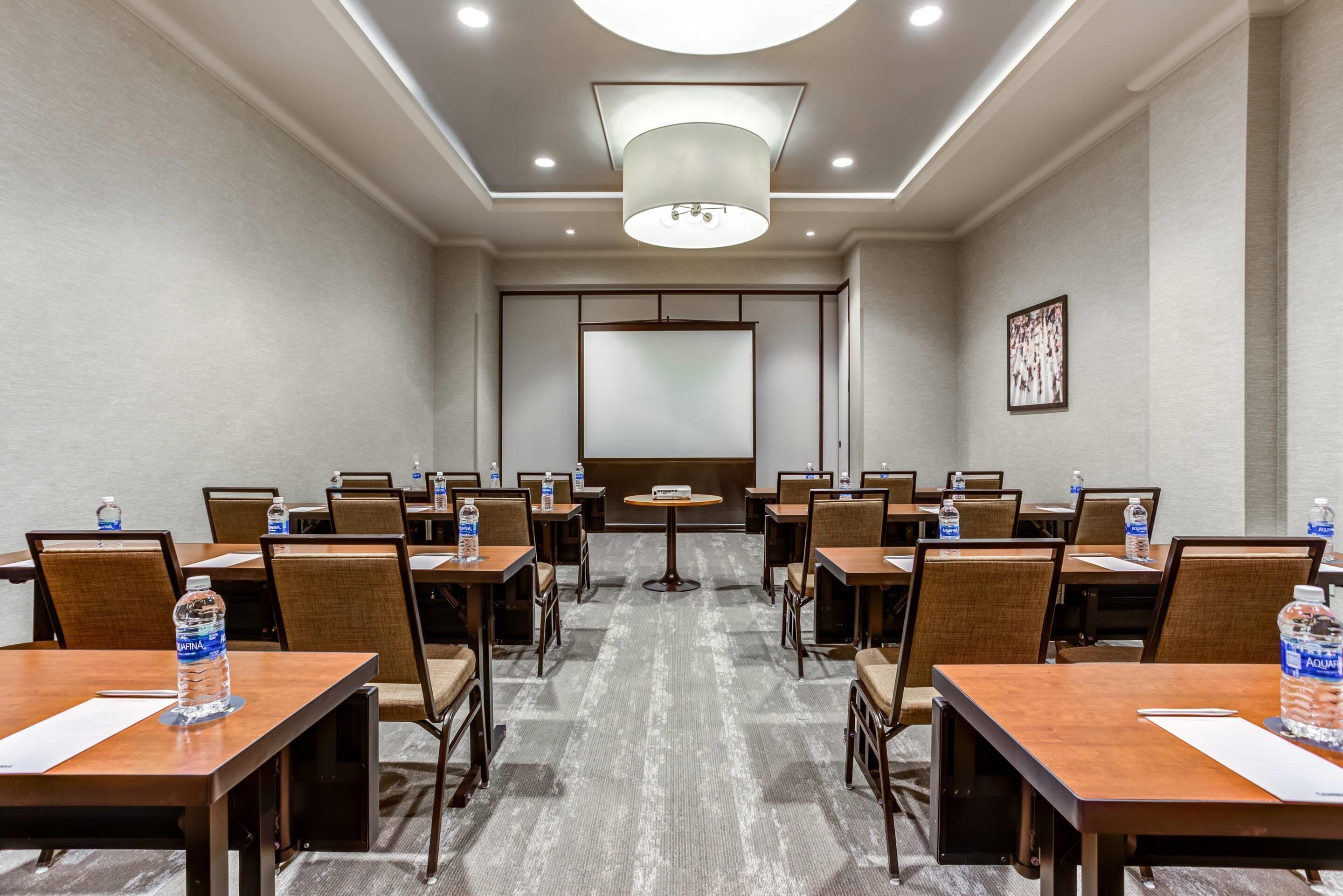 Uma sala de conferências com fileiras de mesas e cadeiras, equipada com garrafas de água, um projetor e uma grande luminária no teto.