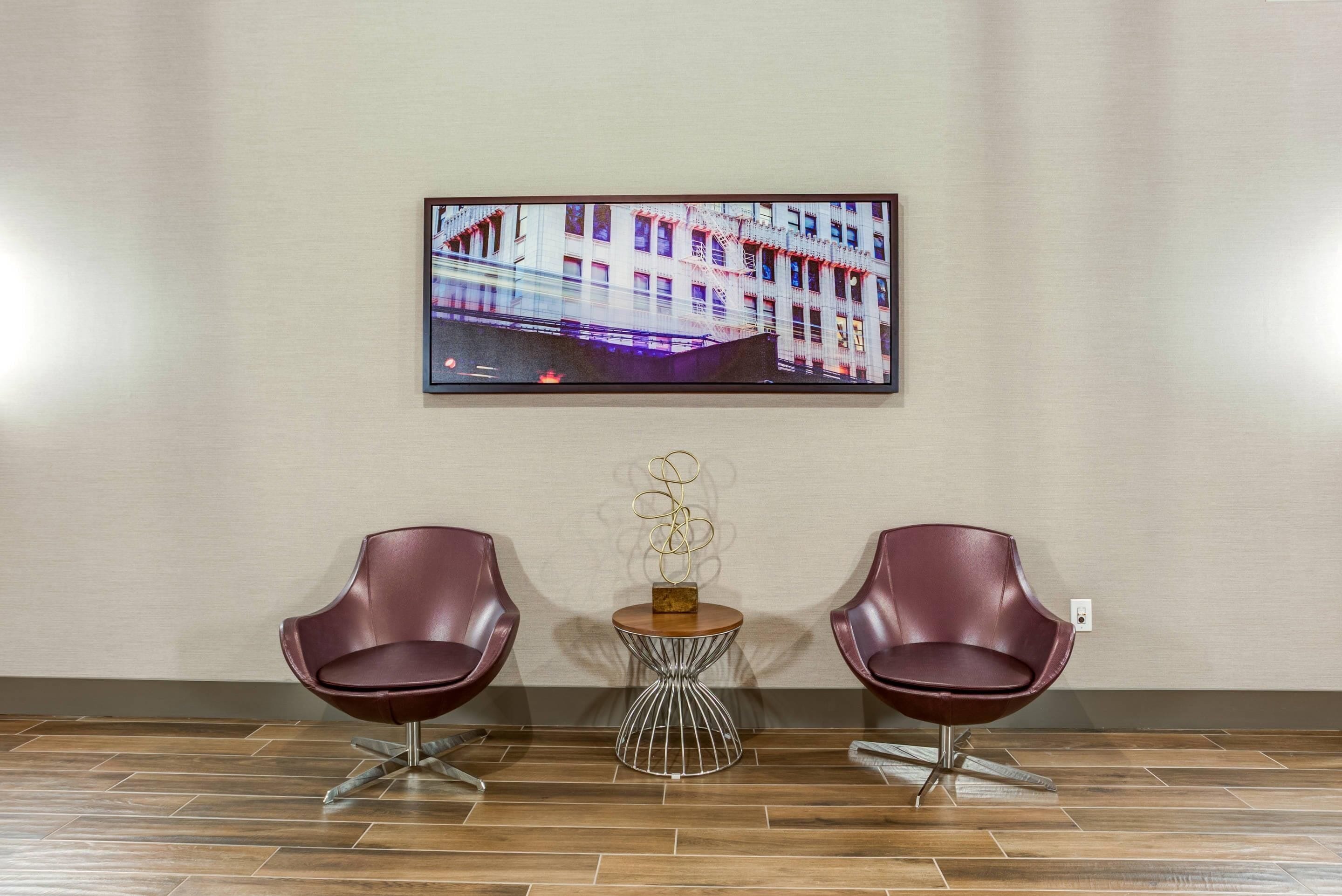 Un'area d'attesa moderna con due poltrone rosse, un tavolino rotondo con un centrotavola decorativo e un quadro di un edificio appeso al muro.