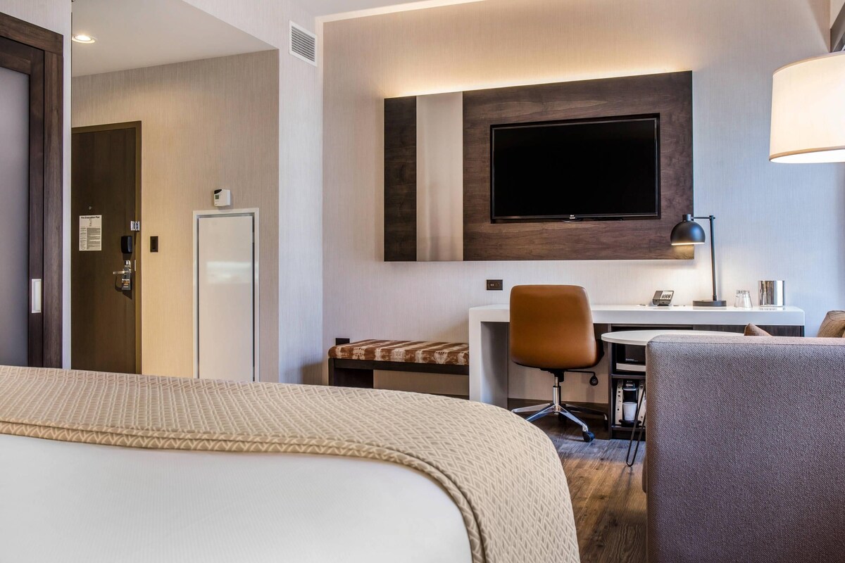 Una habitación de hotel con cama, escritorio, silla, televisor montado en la pared y lámpara.