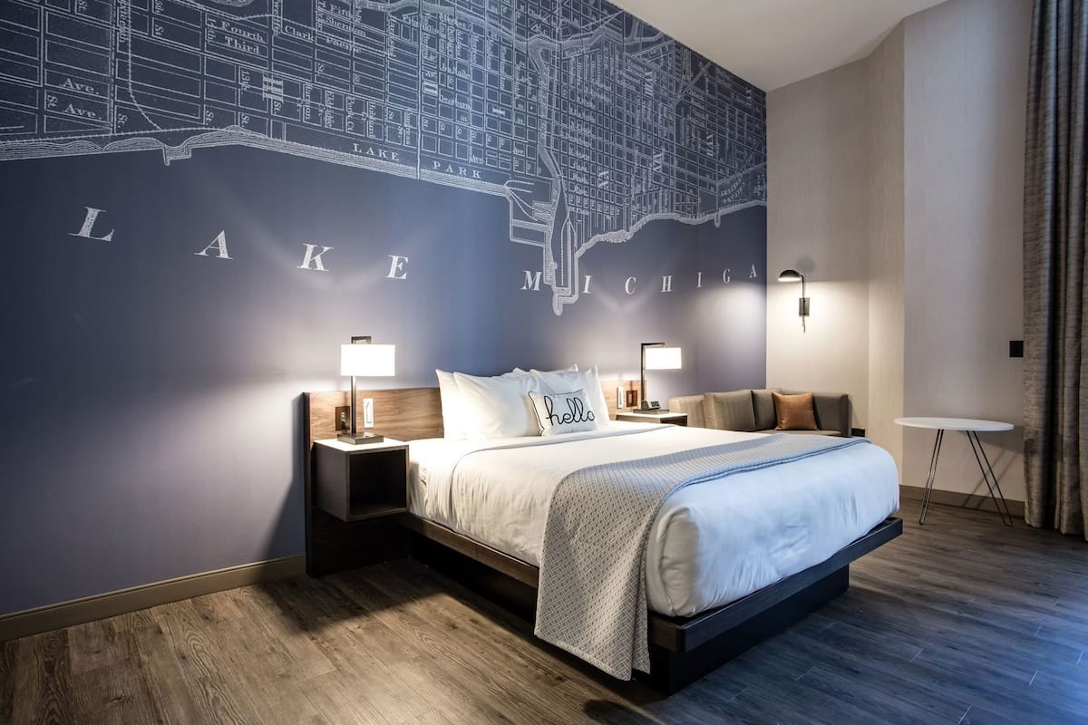 Une chambre d’hôtel avec une grande fresque représentant une carte, un lit avec une tête de lit blanche, deux tables de chevet avec des lampes et une petite table ronde.