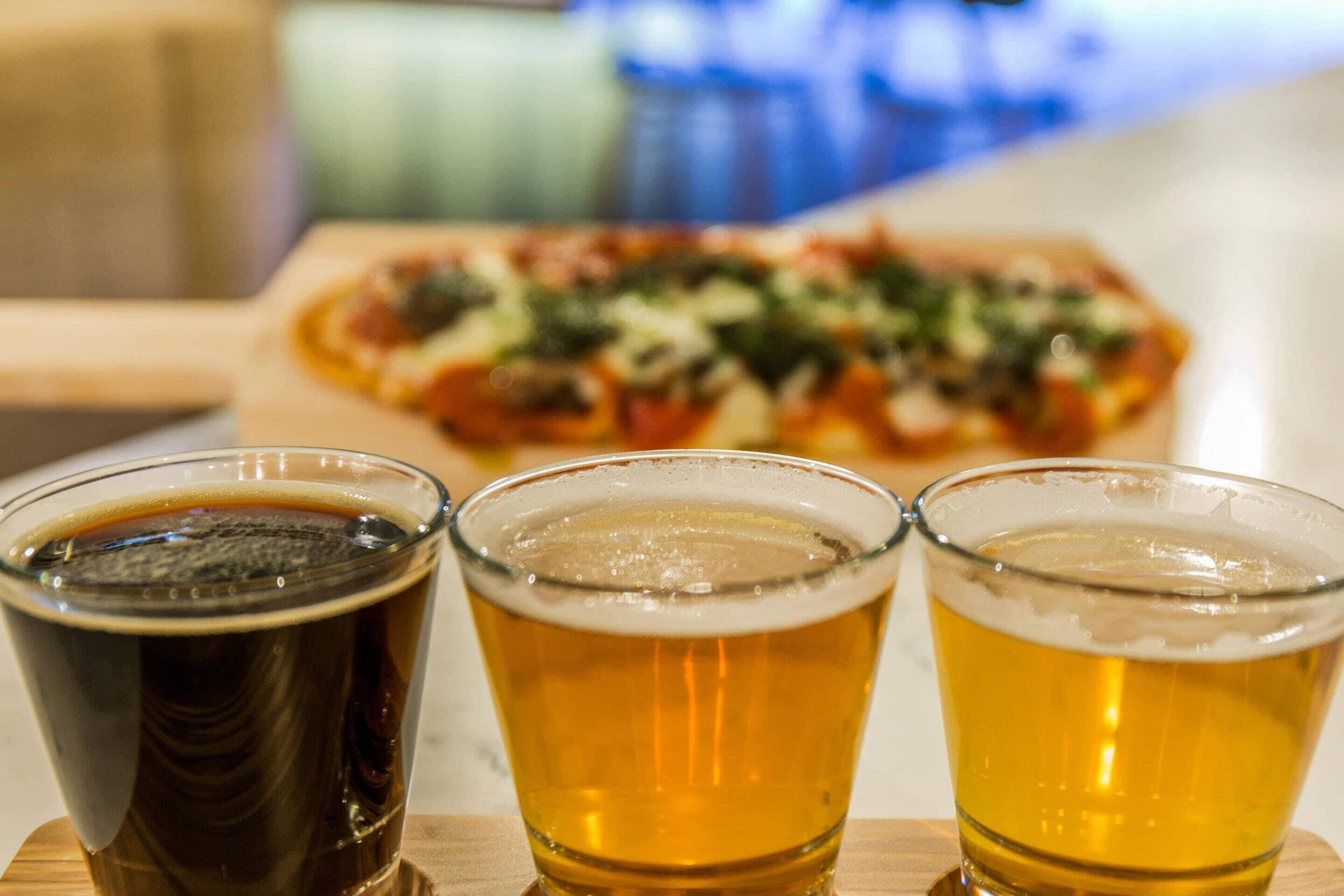 Three glasses of beer on a wooden table with a pizza in the background.