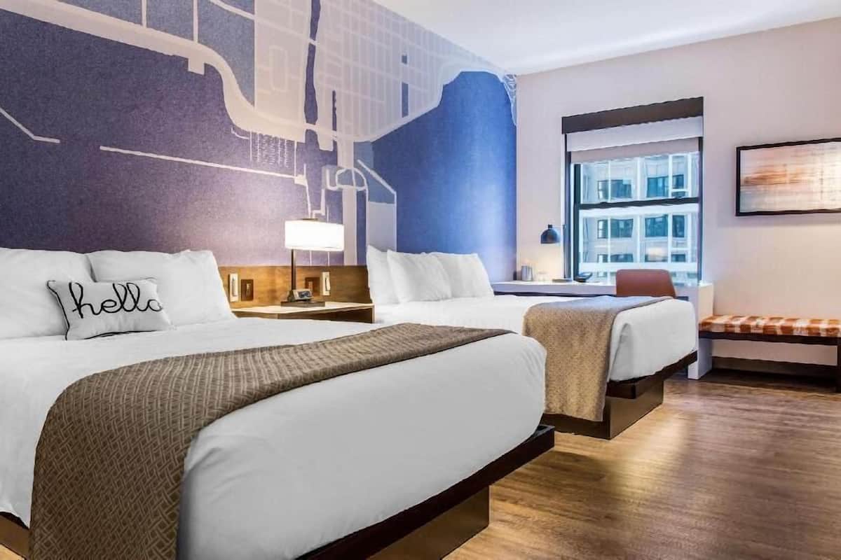 Une chambre d’hôtel avec deux lits, un banc, une fresque représentant un paysage urbain, une fenêtre avec vue et une œuvre d’art encadrée au mur.