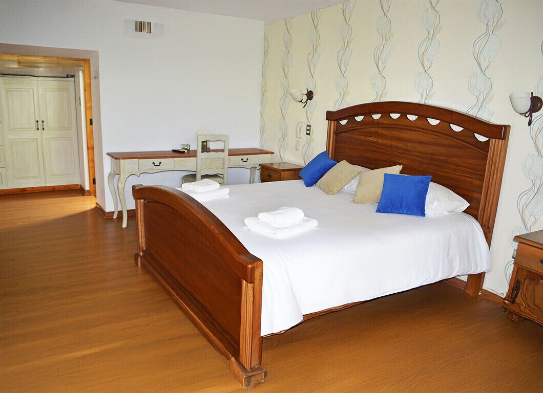 Ein ordentlich bezogenes Bett mit weißen Leinen und blau-beigen Kissen in einem Zimmer mit Holzdielen und einem Schreibtisch mit Stuhl.