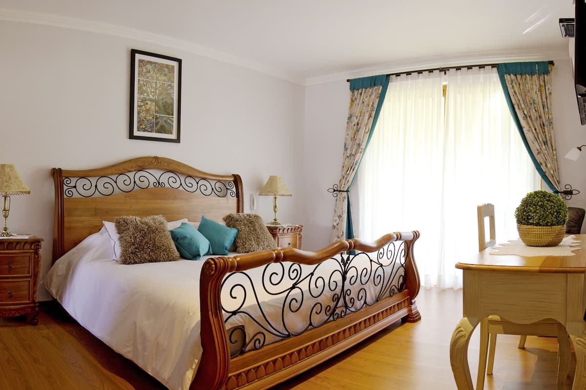 A bedroom with a wooden bed, two bedside tables with lamps, a chair, a desk, and a window with curtains.