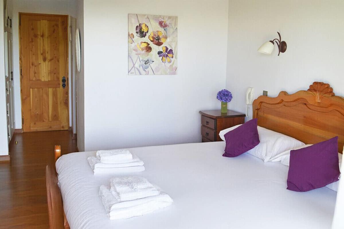 A bedroom with a wooden door, a bed with white linens and purple pillows, a nightstand with a vase of flowers, and a wall-mounted lamp.