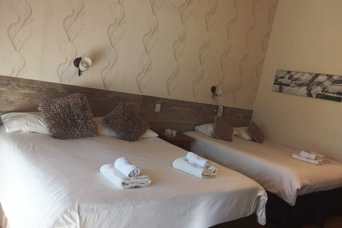 A hotel room with two beds, each with a white bedspread and pillows, a wooden headboard, and a wall-mounted light fixture.