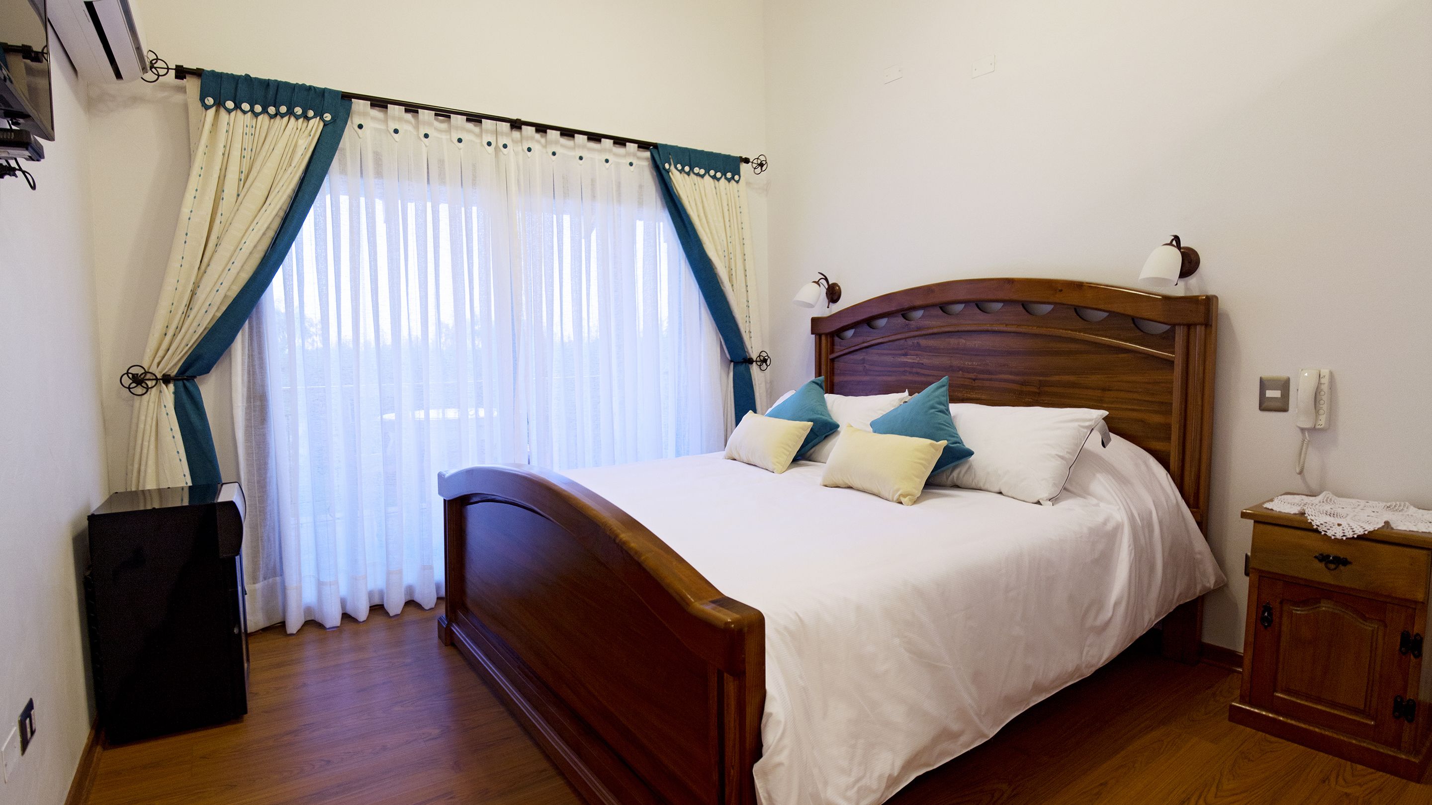 A neatly made bed with a wooden headboard, a bedside table with a lamp, and a window with curtains.