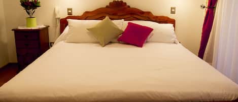 Una cama bien hecha con ropa blanca y dos almohadas, un cabecero de madera, una mesita de noche con una lámpara y una planta en maceta.