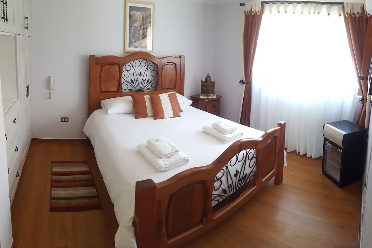 A neatly made bed with white linens and brown pillows, a wooden headboard with a decorative pattern, a small bedside table with a lamp, and a window with curtains.