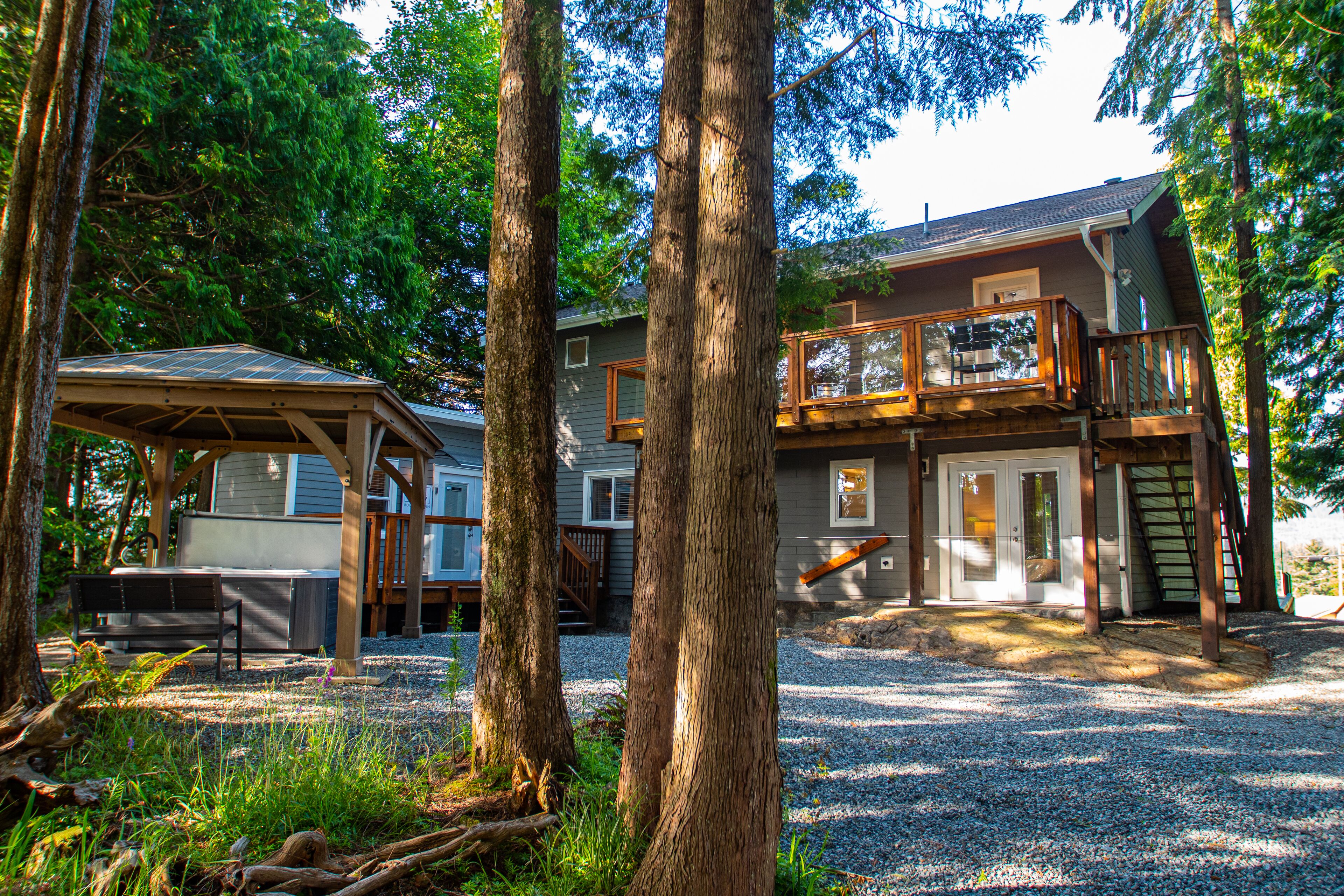 A two-story house with a deck, surrounded by trees and a gravel driveway.