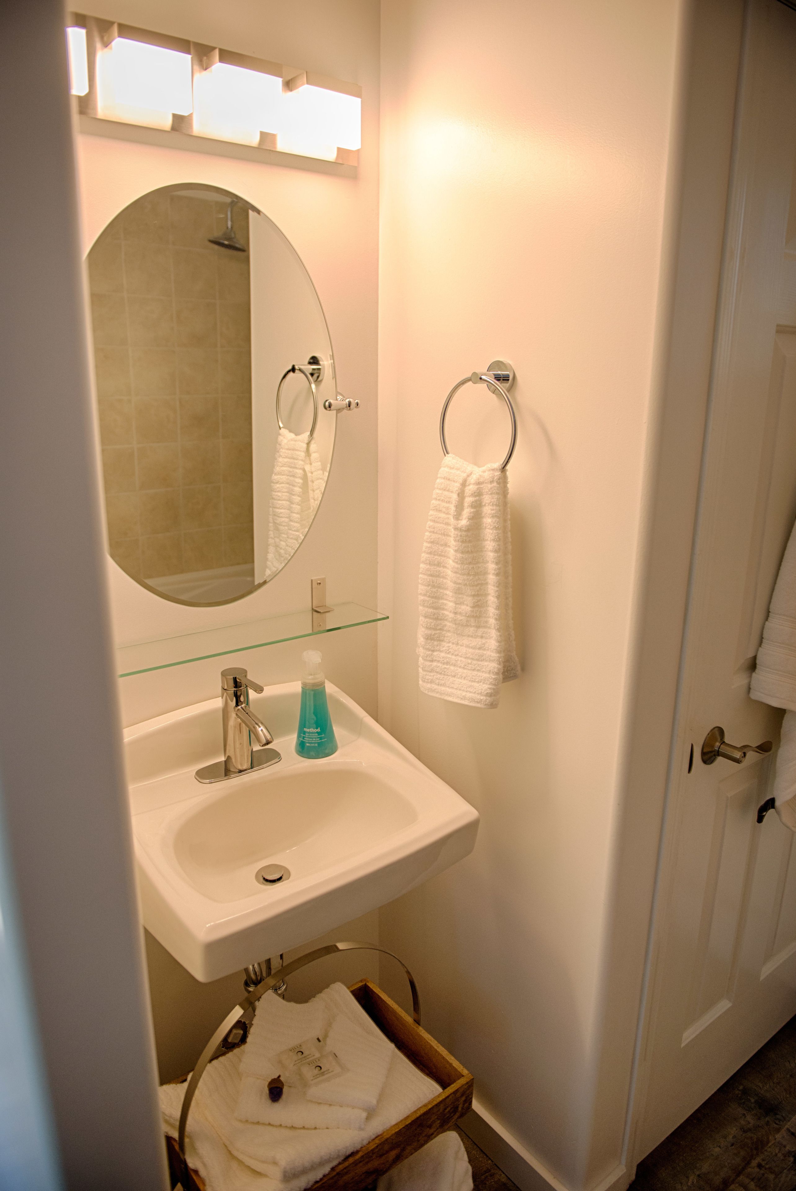 A bathroom with a white sink, a glass shelf, a mirror, a towel rack, and a wooden basket.