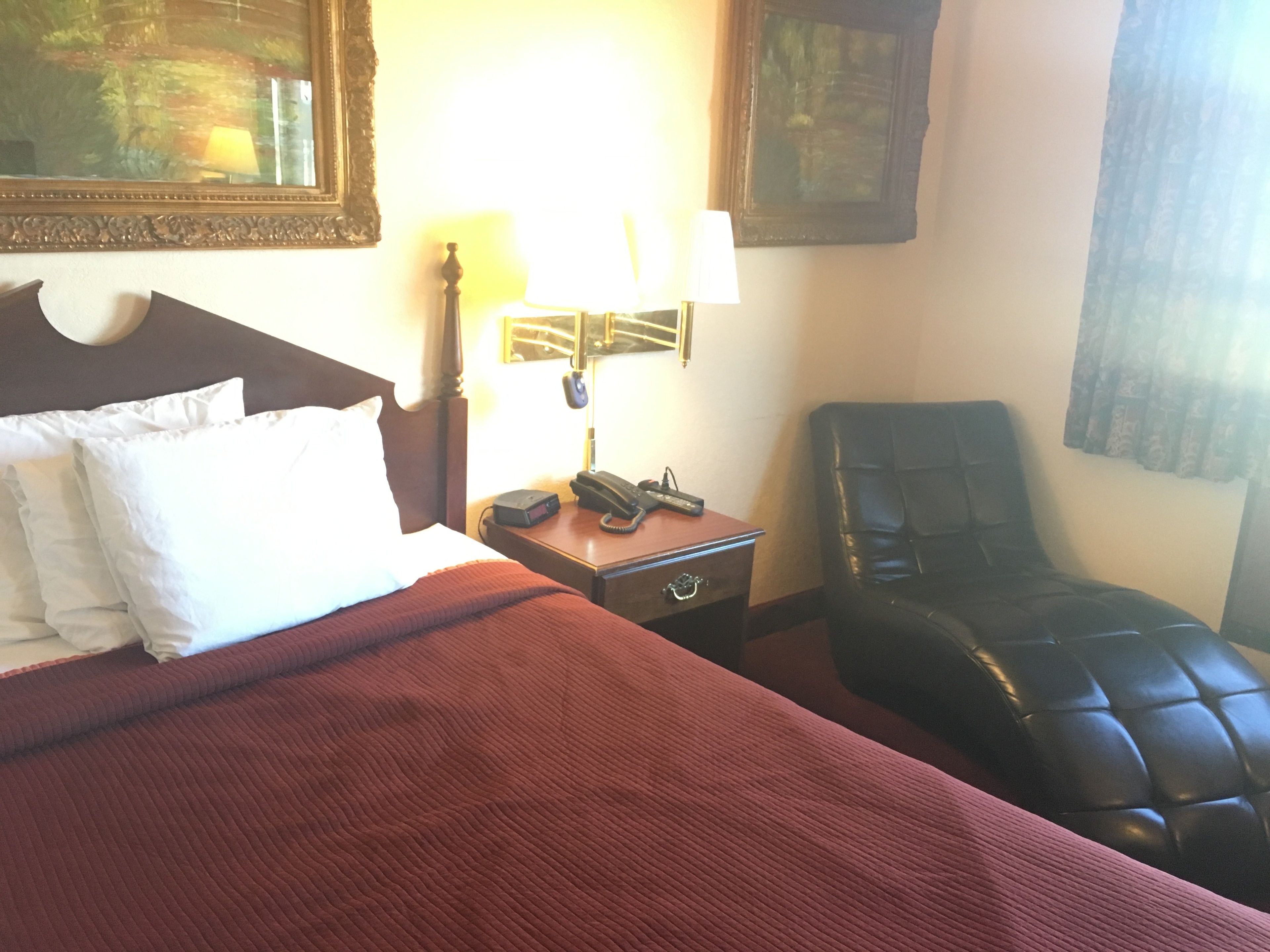 Quarto de hotel com cama, mesa de cabeceira com abajur, cadeira e escrivaninha com telefone.