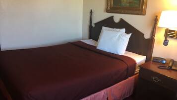 Un letto rifatto con una coperta rosso scuro e cuscini bianchi, un comodino in legno con una lampada, e un quadro appeso al muro.