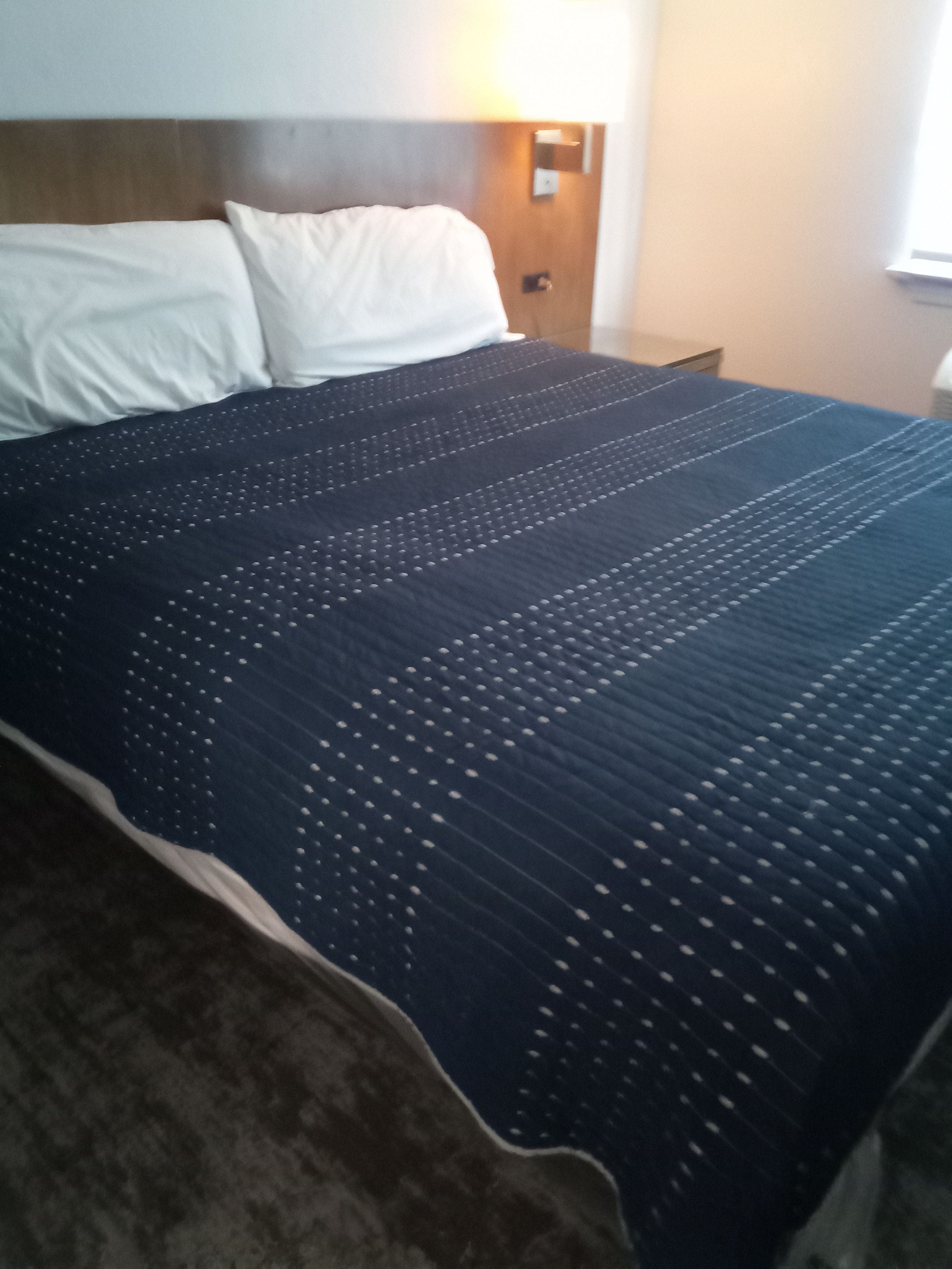 Una cama con colcha oscura, almohadas blancas, cabecera de madera y una lámpara fijada a la pared.
