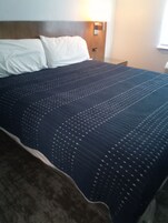 Una cama con colcha oscura, almohadas blancas, cabecera de madera y una lámpara fijada a la pared.