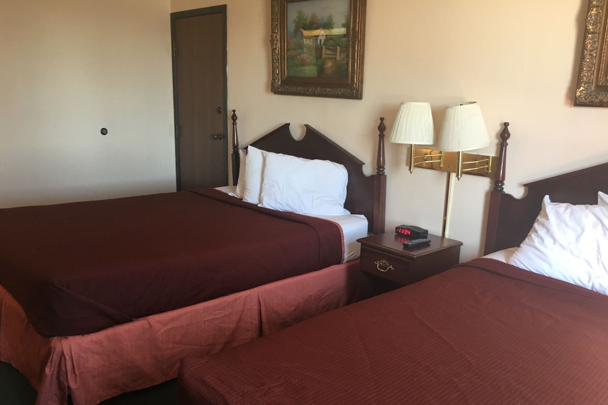 A hotel room with two beds, a nightstand with a clock, a lamp, and a painting on the wall.