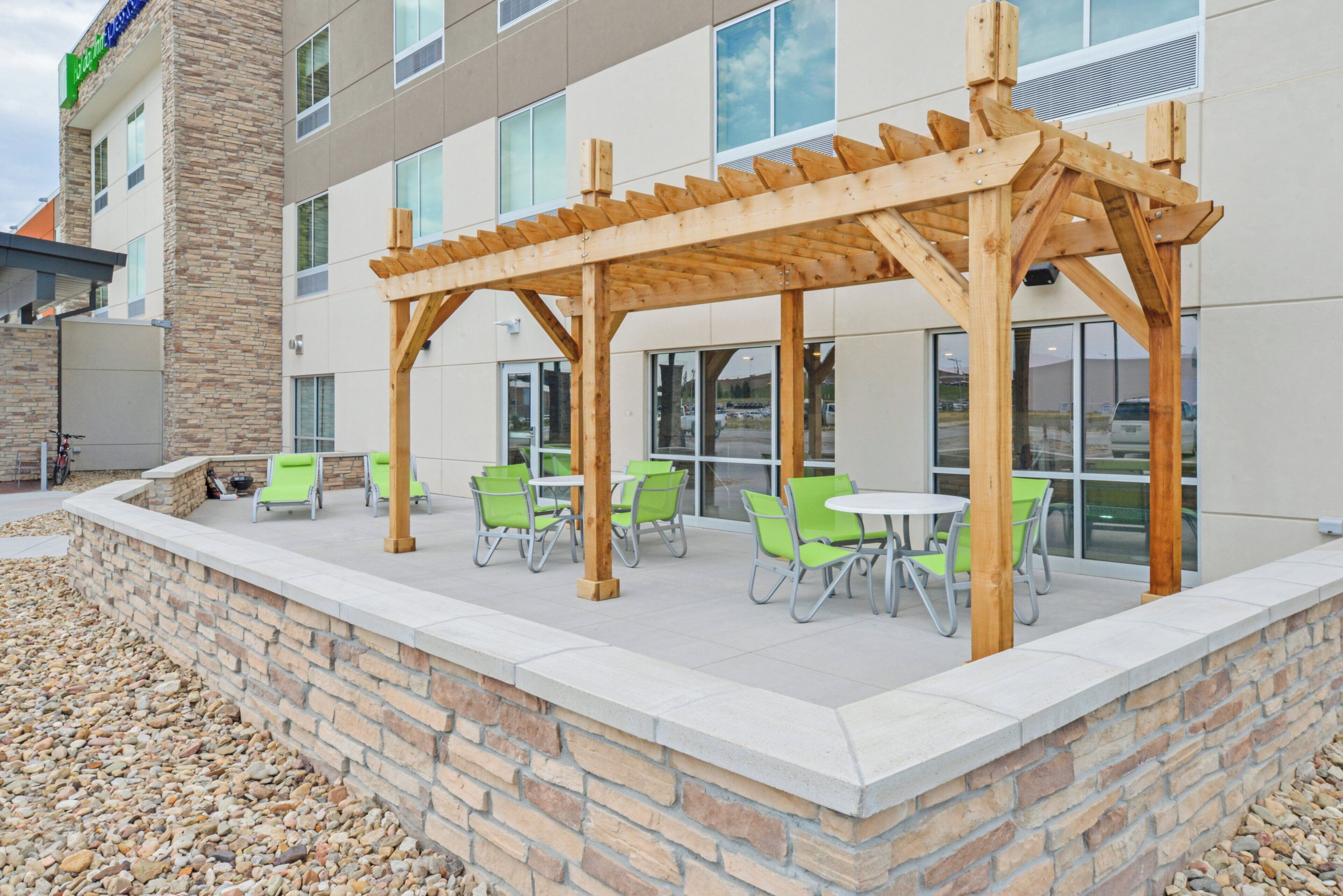 An outdoor patio area with wooden pergola, green chairs, and tables, adjacent to a building with large windows.