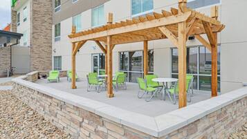 An outdoor patio area with wooden pergola, green chairs, and tables, adjacent to a building with large windows.