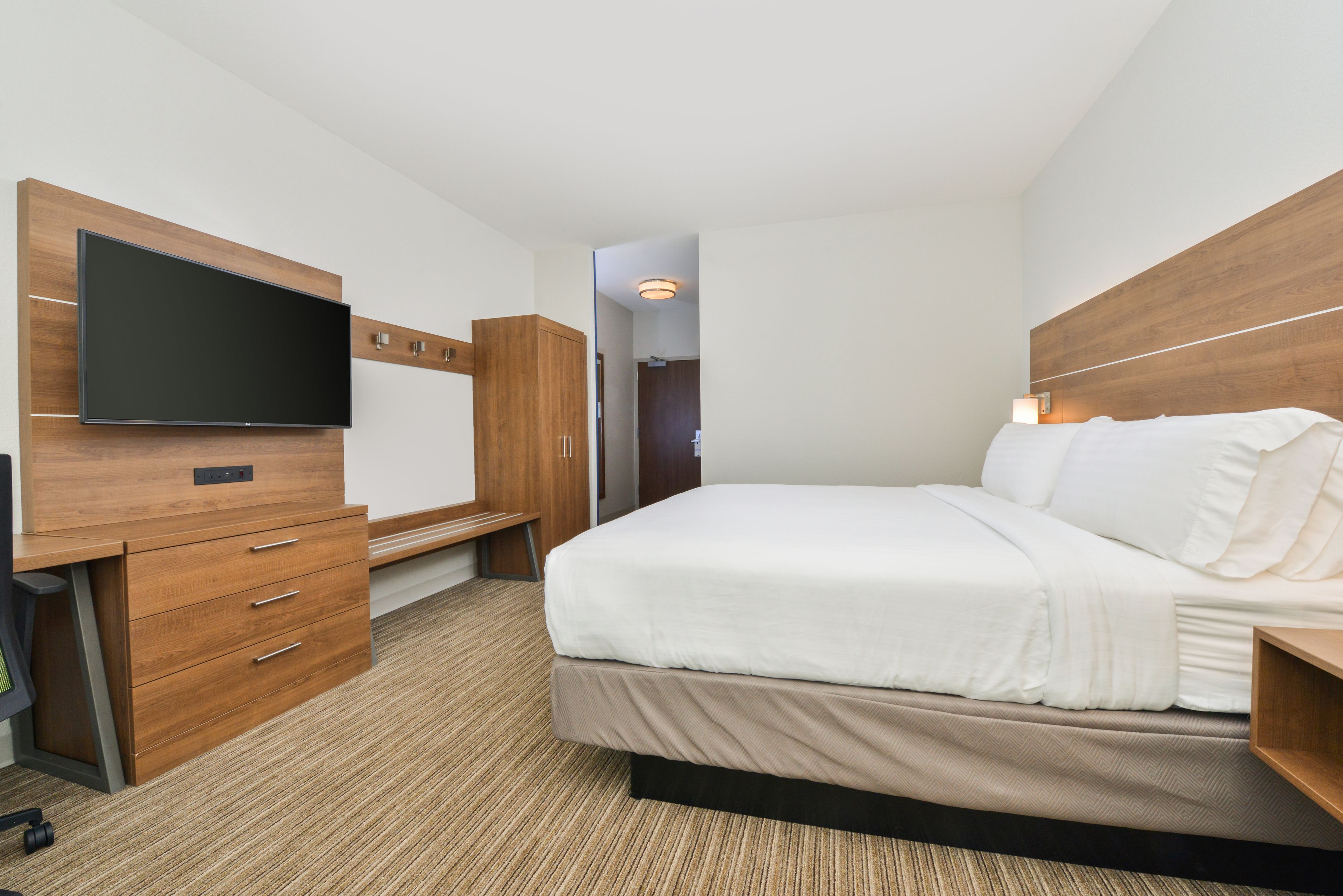 Quarto de hotel com cama, televisão presa a um móvel de madeira e escrivaninha com gavetas.