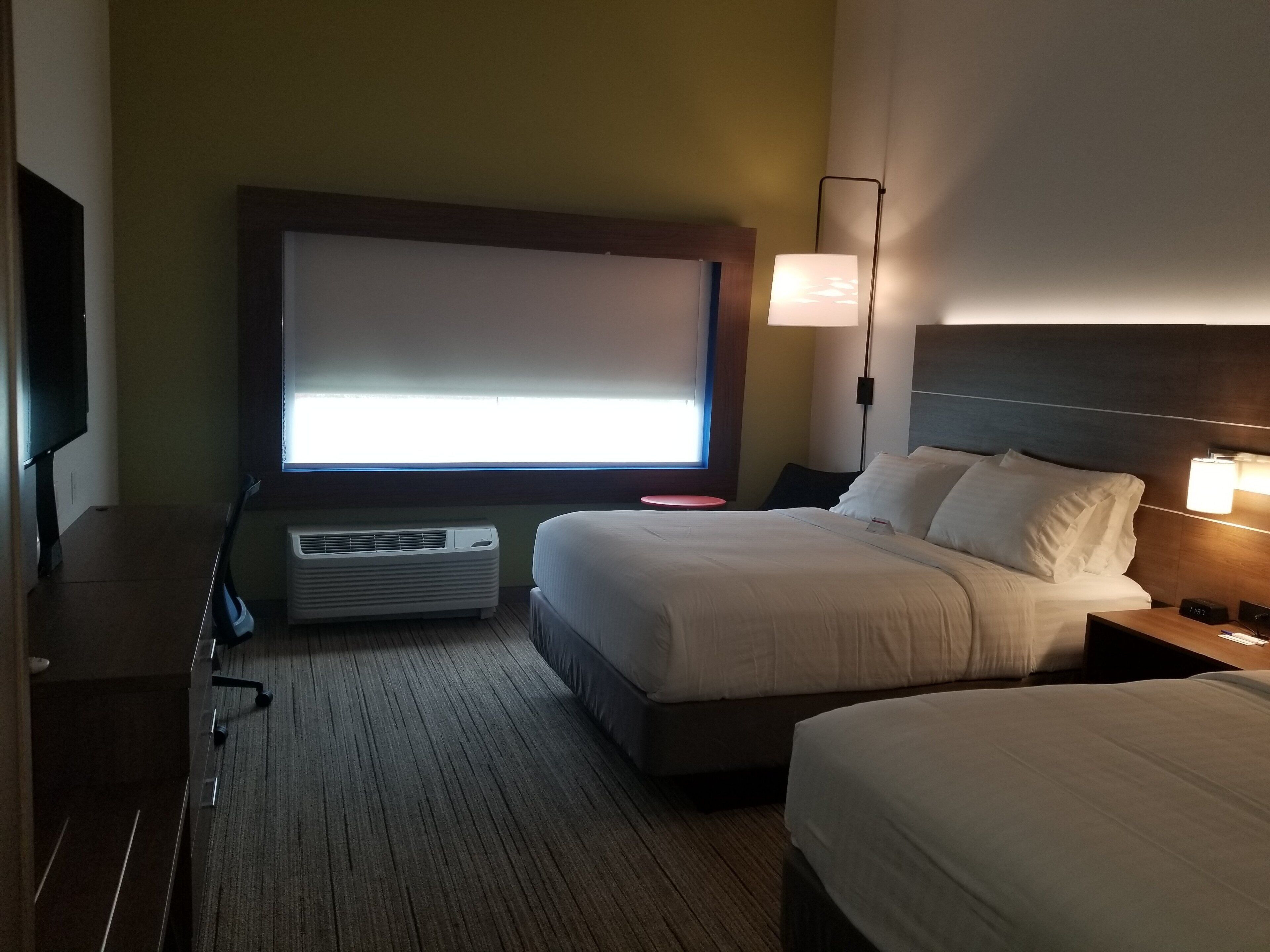 A hotel room with a bed, bedside table, lamp, air conditioning unit, and a television.