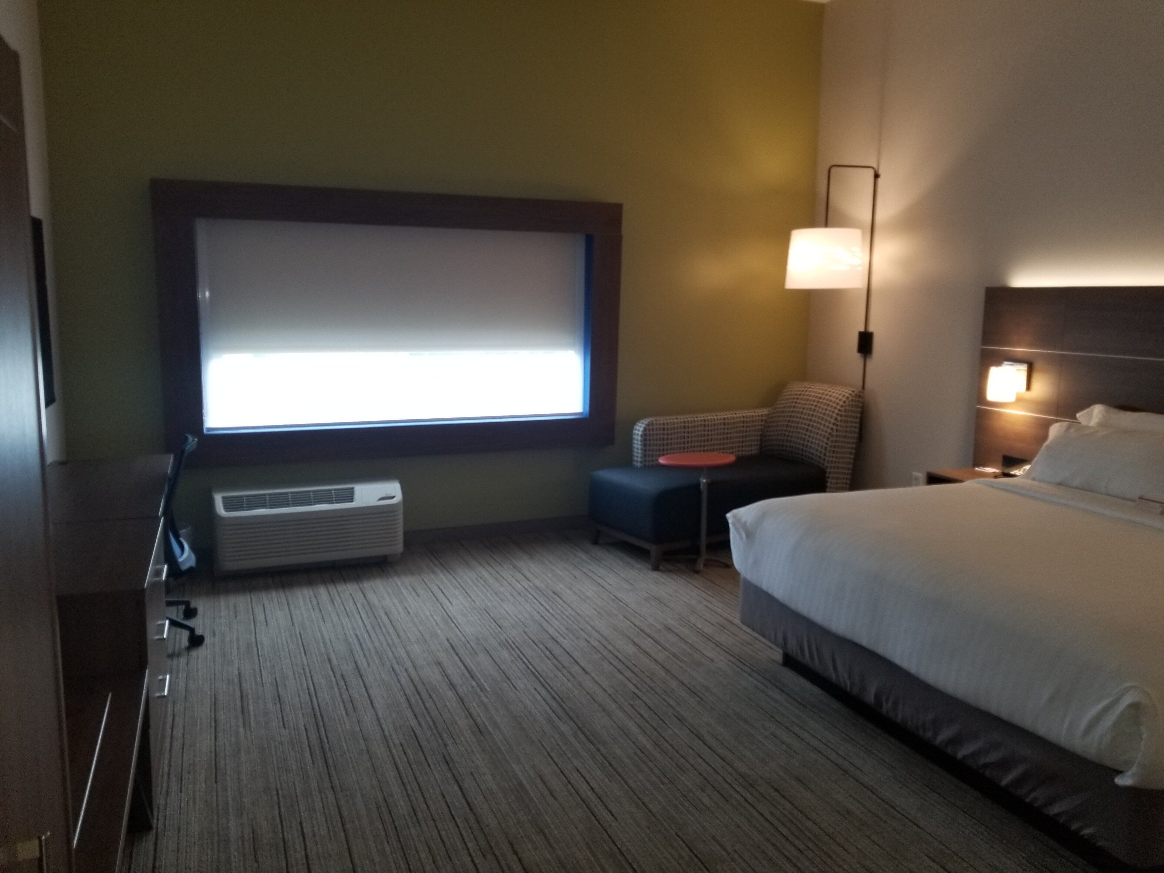 A hotel room with a bed, a desk, a chair, a window with blinds, a lamp, and an air conditioning unit.