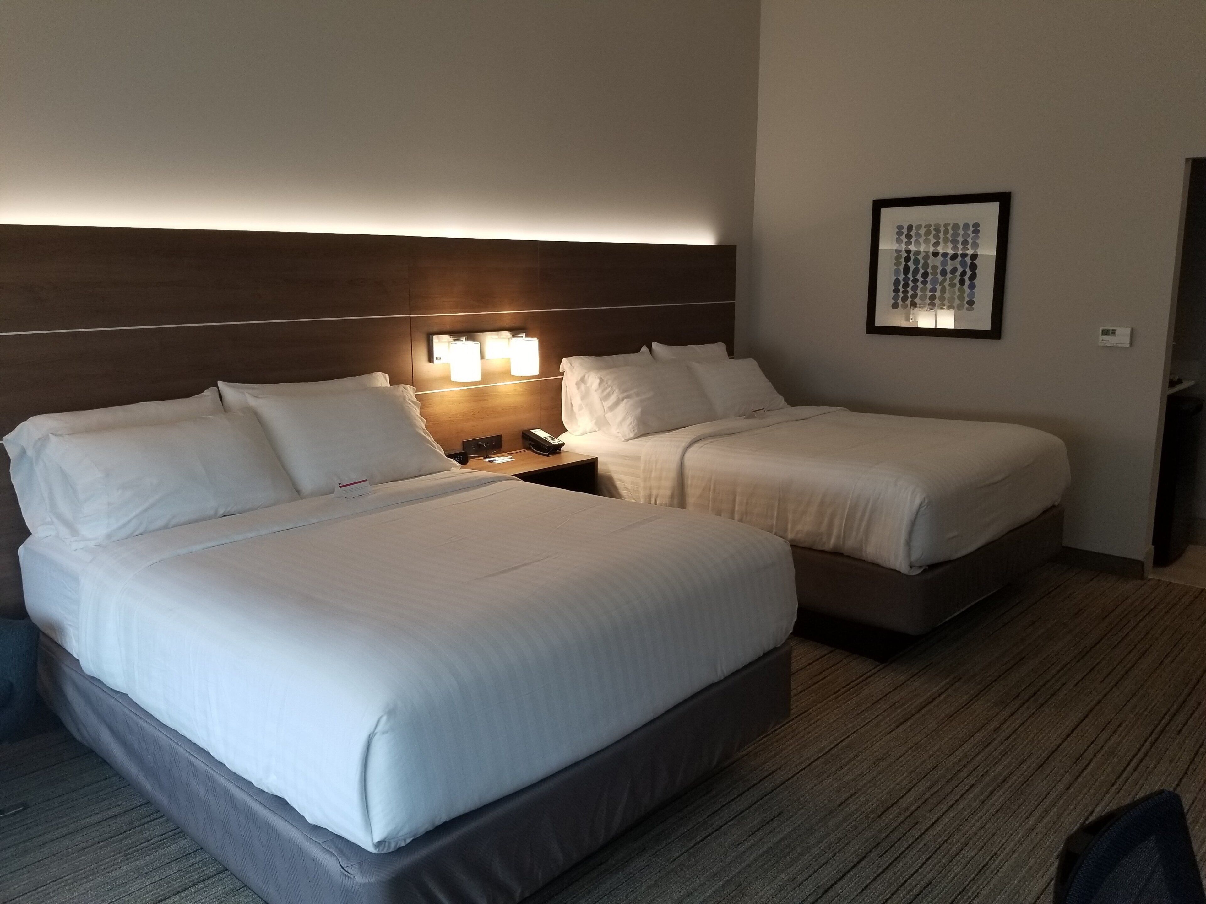 A hotel room with a large bed, two smaller beds, a nightstand with a lamp, and a framed picture on the wall.