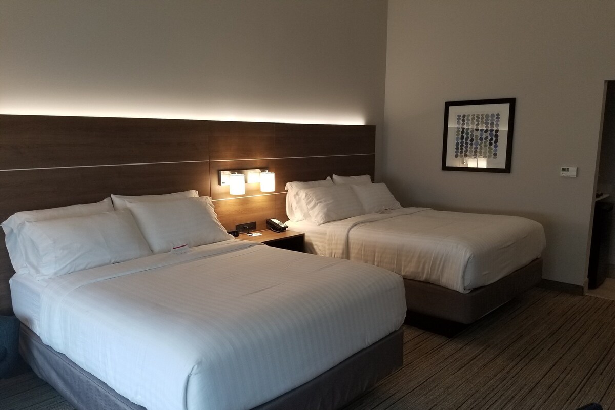 Habitación de hotel con una cama grande, dos camas más pequeñas, una mesita de noche con una lámpara y un cuadro en la pared.