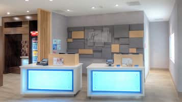 Ein Hotelrezeptionstresen mit einem digitalen Display, einem Coca-Cola-Kühlschrank und einer Wand mit geometrischem Muster.