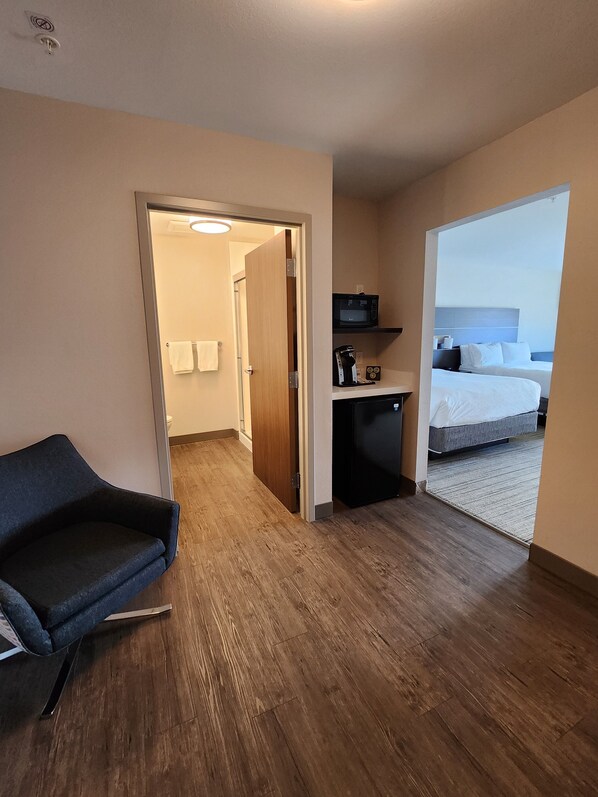 Habitación de hotel con piso de madera, cama, kitchenette pequeña con microondas y zona de estar con una silla.