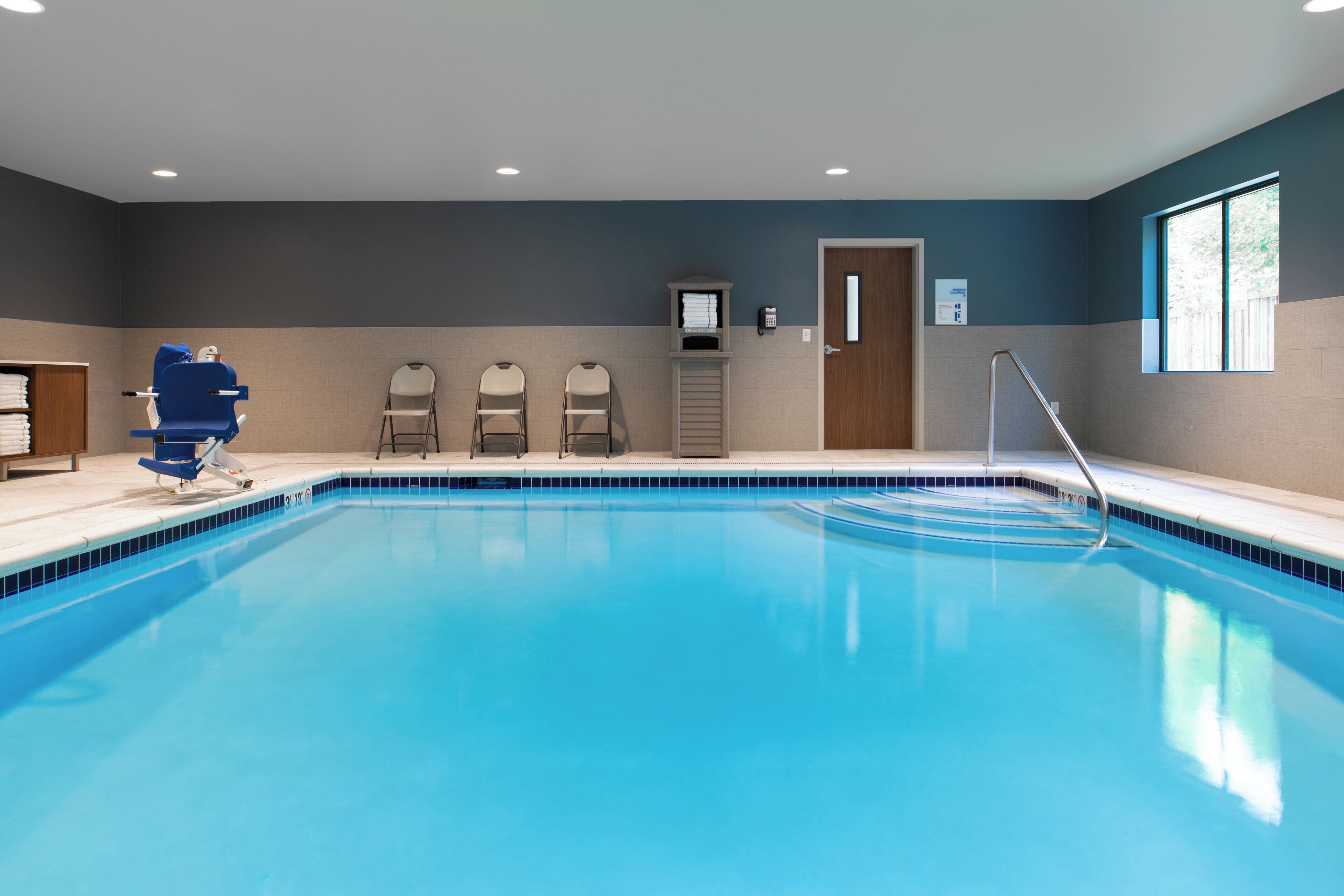 A swimming pool with a blue tiled edge, a changing room with a blue chair, and a wooden door.