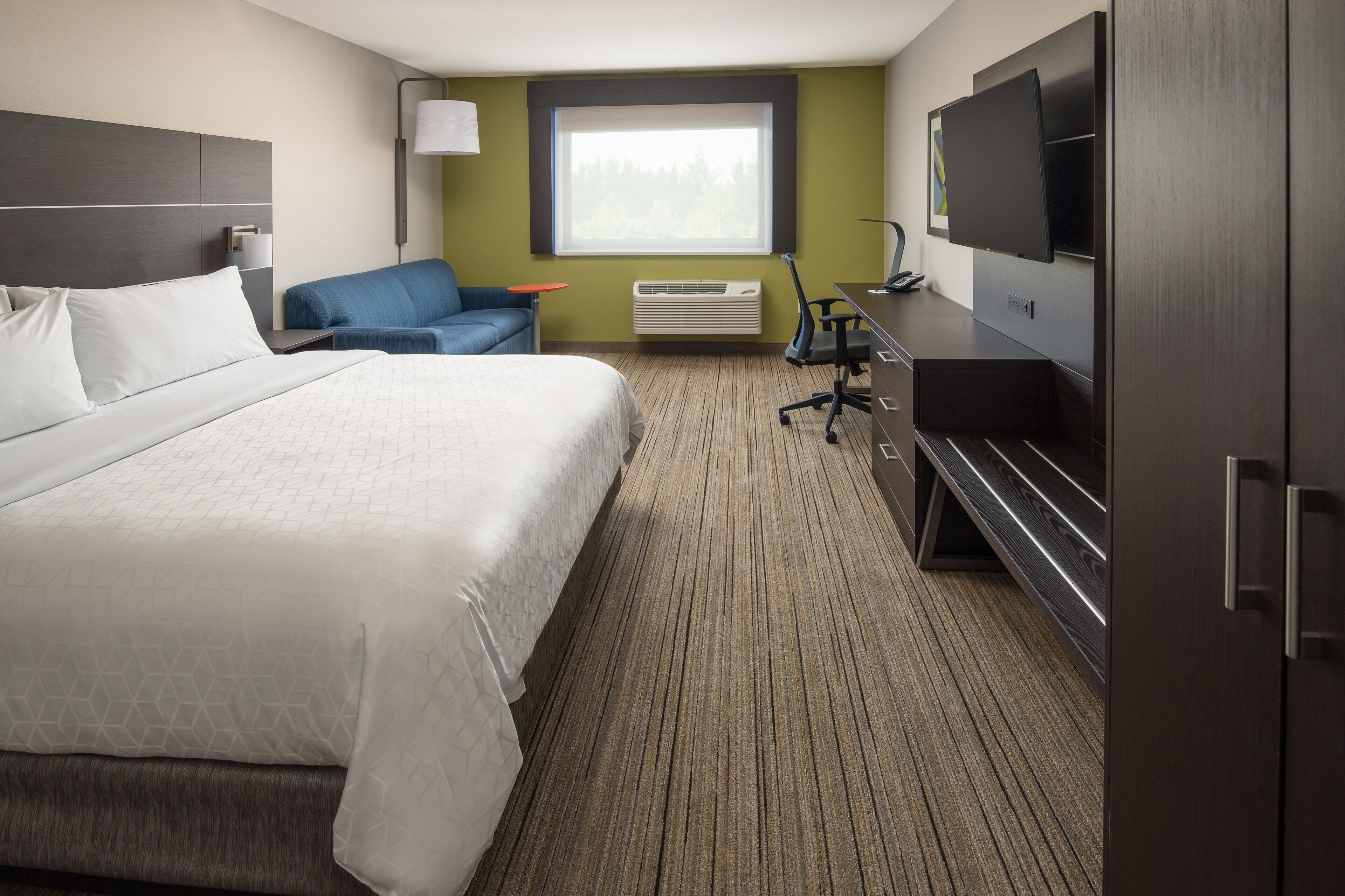 A hotel room with a large bed, a desk with a flat-screen TV, a chair, and a window with a view of trees.