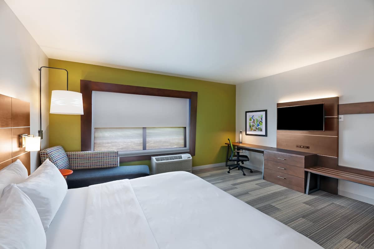 A hotel room with a large bed, a desk, a chair, a flat-screen TV, and a window with blinds.