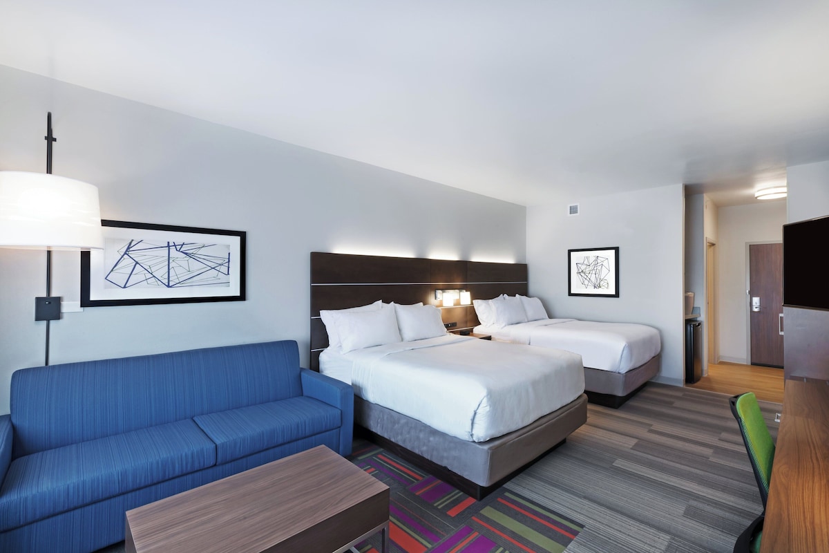 Quarto de hotel com um sofá azul, duas camas, uma mesa de madeira e obras de arte nas paredes.