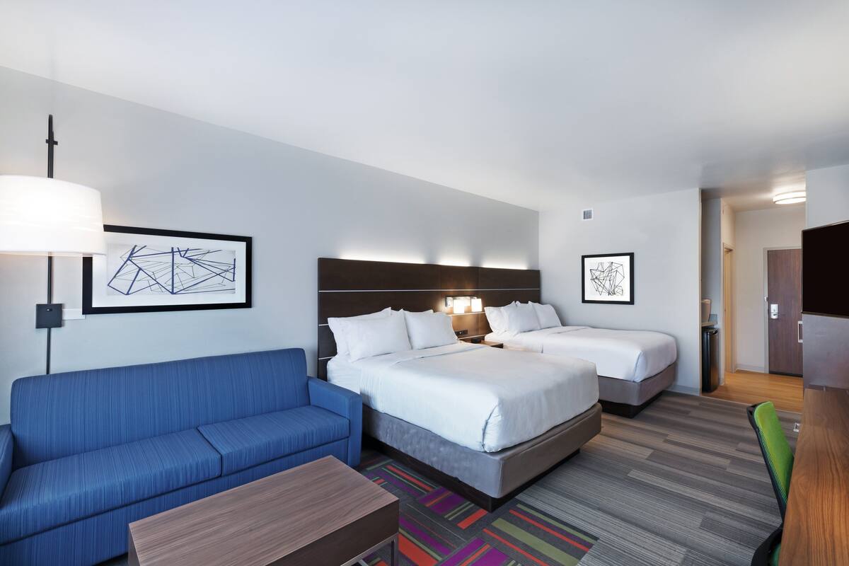 A hotel room with a blue sofa, two beds, a wooden table, and artwork on the walls.