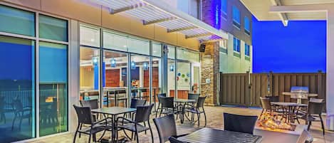 An outdoor patio area with tables and chairs, a barbecue grill, and a view of a building with large windows and a stone accent wall.