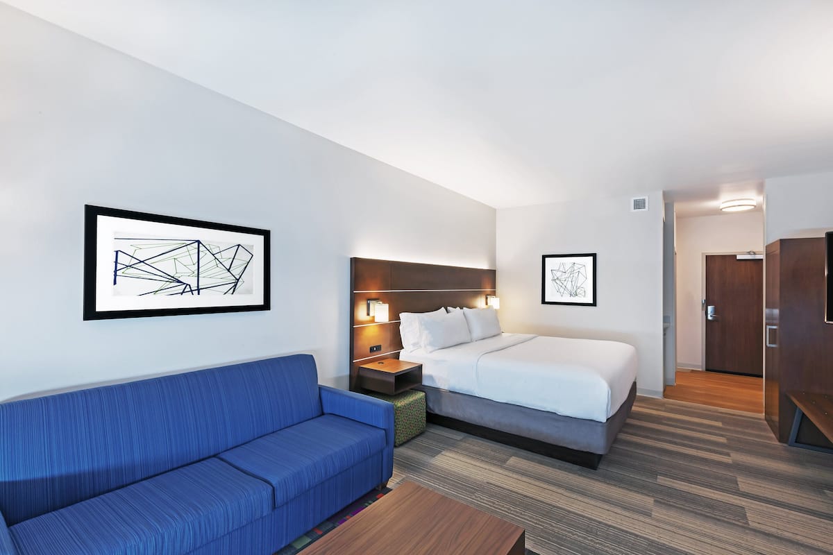 A hotel room with a blue sofa, a bed with white bedding, a nightstand with a lamp, and two framed abstract artworks on the walls.