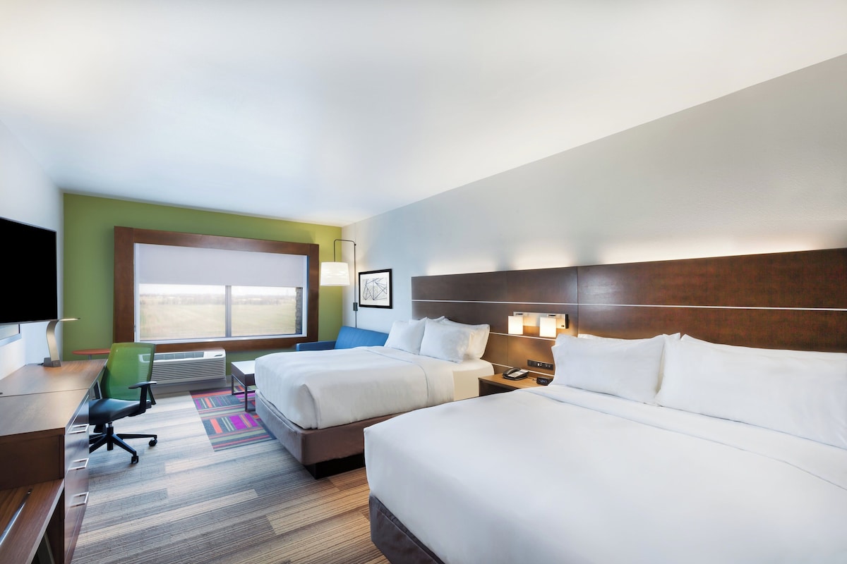 Quarto de hotel com duas camas, uma escrivaninha, uma cadeira, uma televisão e uma janela grande.