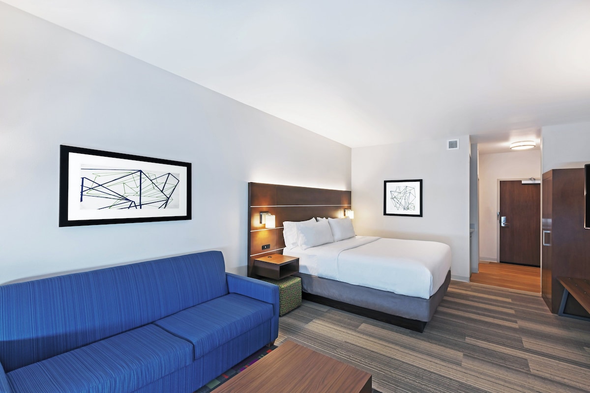 Quarto de hotel com sofá azul, cama com roupa de cama branca, mesa de cabeceira com abajur e duas obras de arte abstratas emolduradas nas paredes.