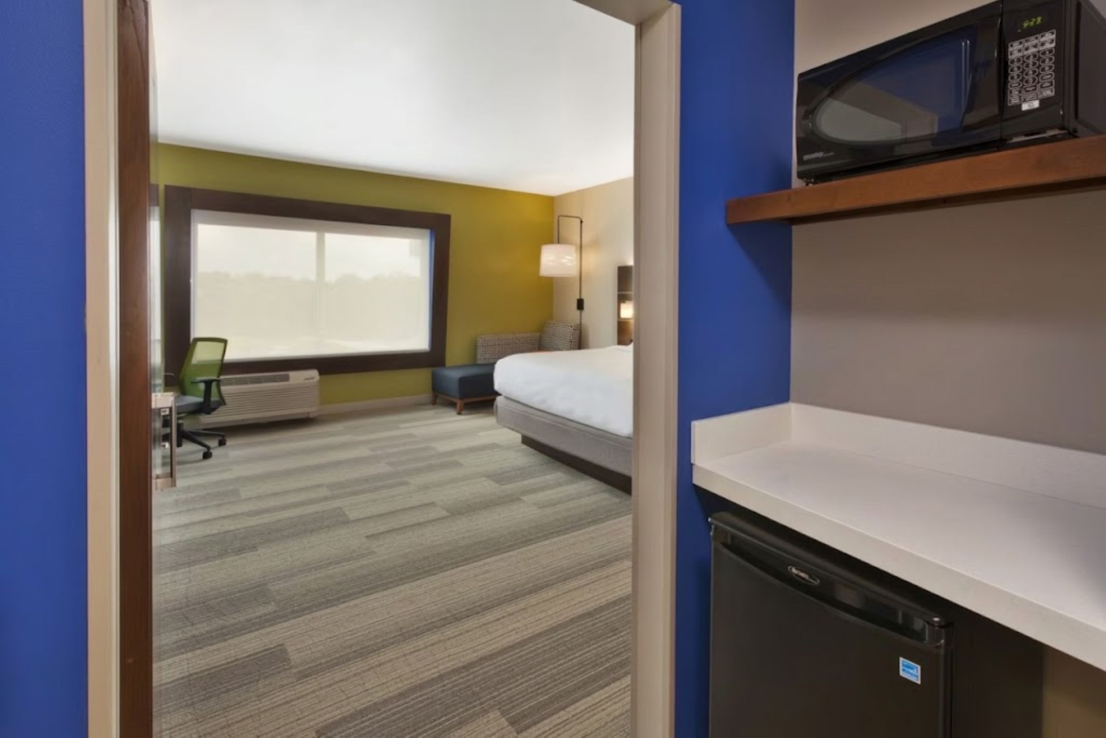 A hotel room with a bed, a desk, a chair, a microwave, and a small refrigerator.