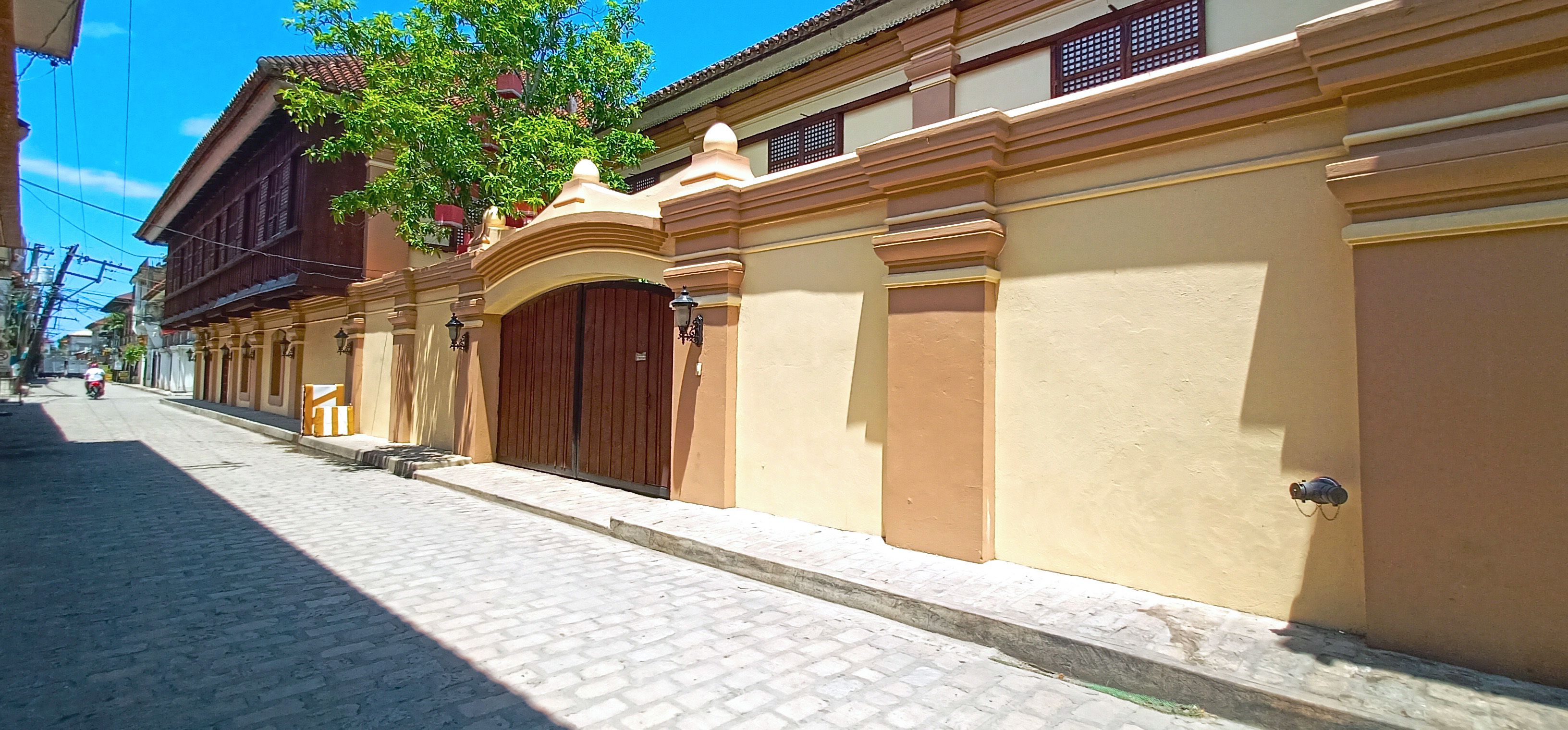 Calle empedrada bordeada de edificios con arquitectura tradicional, una gran puerta de madera y un árbol que ofrece sombra.