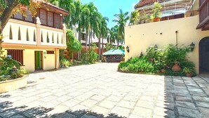 Un patio con un camino empedrado, rodeado de edificios de arquitectura tradicional, plantas en macetas y una pequeña estructura con techo verde.