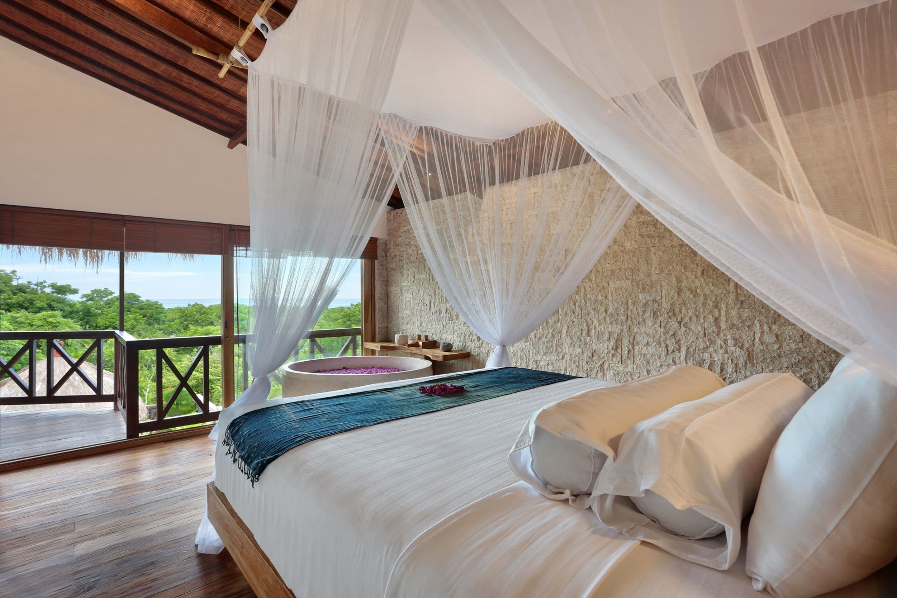 A bedroom with a large bed, white bedding, a view of the outdoors, and a balcony with a wooden railing.