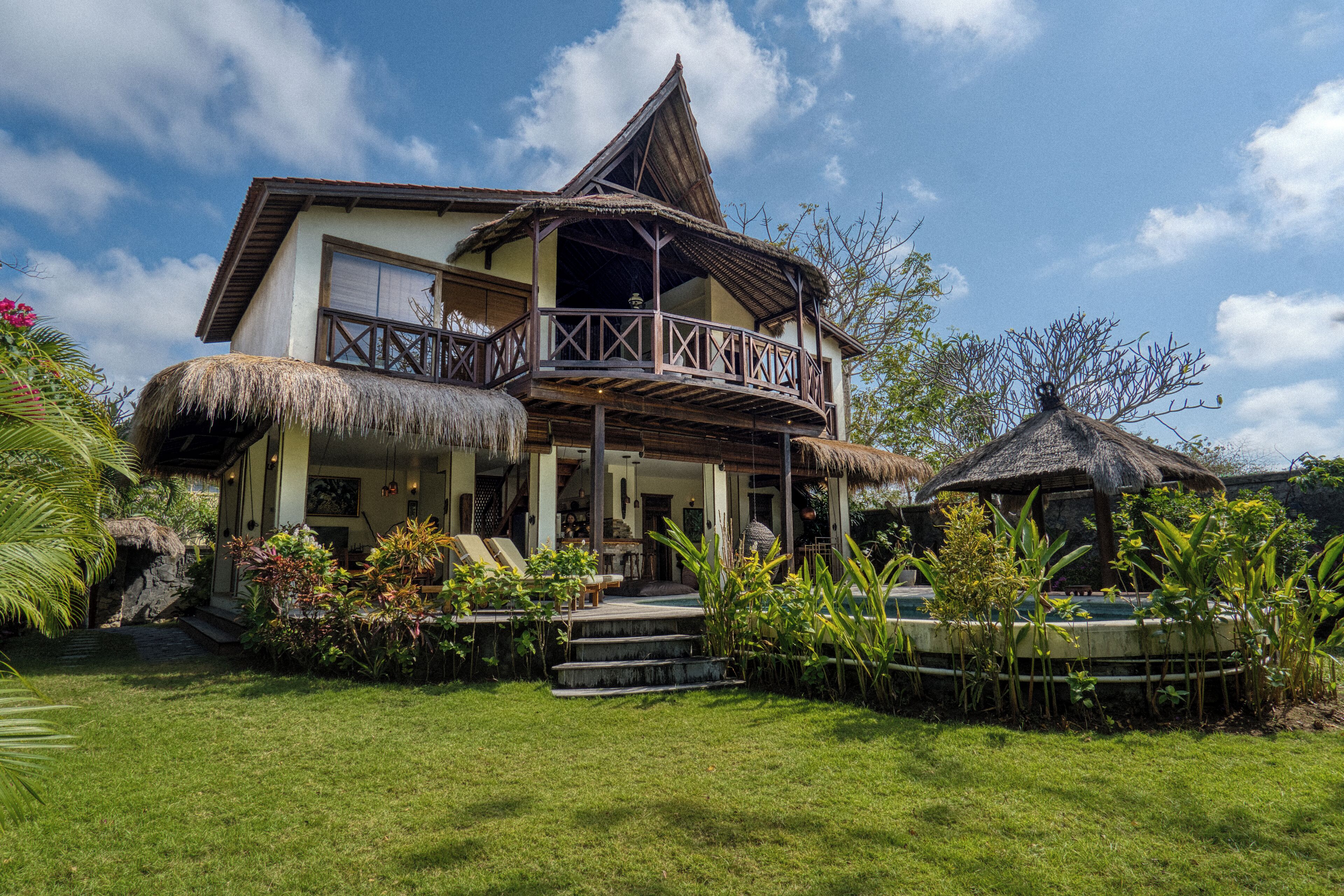 A two-story house with a thatched roof, a balcony, and a garden with tropical plants.