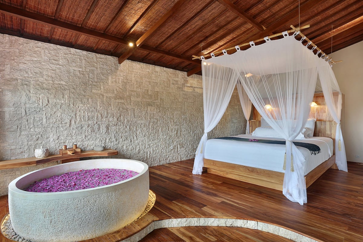 A bedroom with a stone wall, a wooden ceiling, a bed with a canopy, and a stone basin filled with flowers.