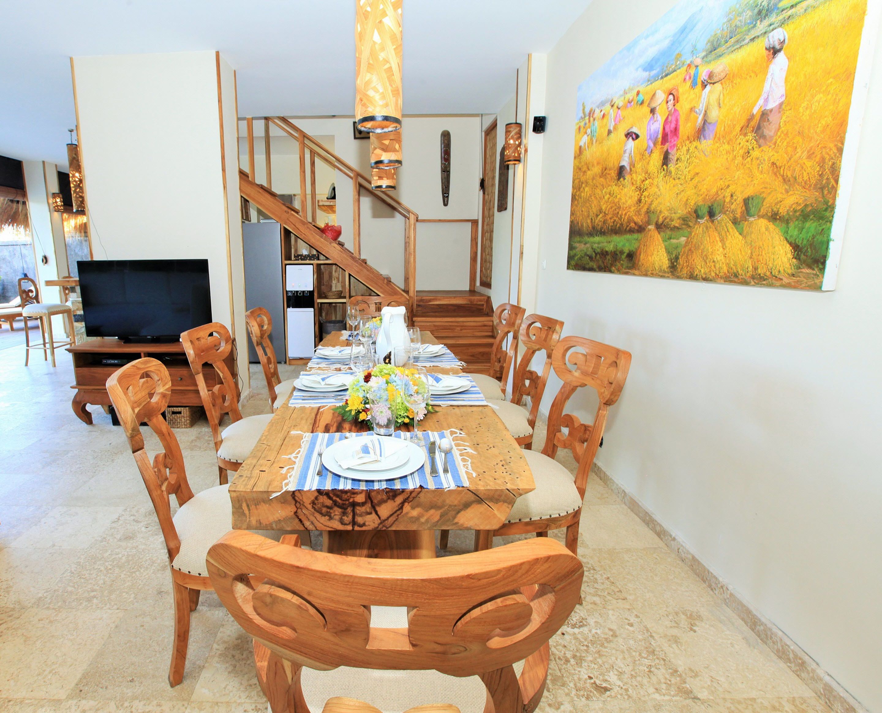 A dining area with a wooden table set for six, surrounded by wooden chairs with cushioned seats. A painting of a field with people is on the wall to the right.