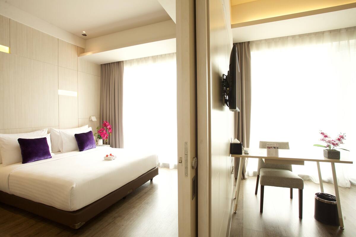 Ein Hotelzimmer mit einem großen Bett, einem Schreibtisch mit Stuhl, einem an der Wand befestigten Fernseher und einem Fenster mit Vorhängen.