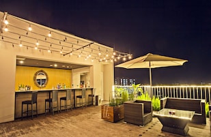 Um bar no terraço com uma parede amarela, balcão, banquetas, mesa de vidro, mobiliário de vime e luzes de corda.