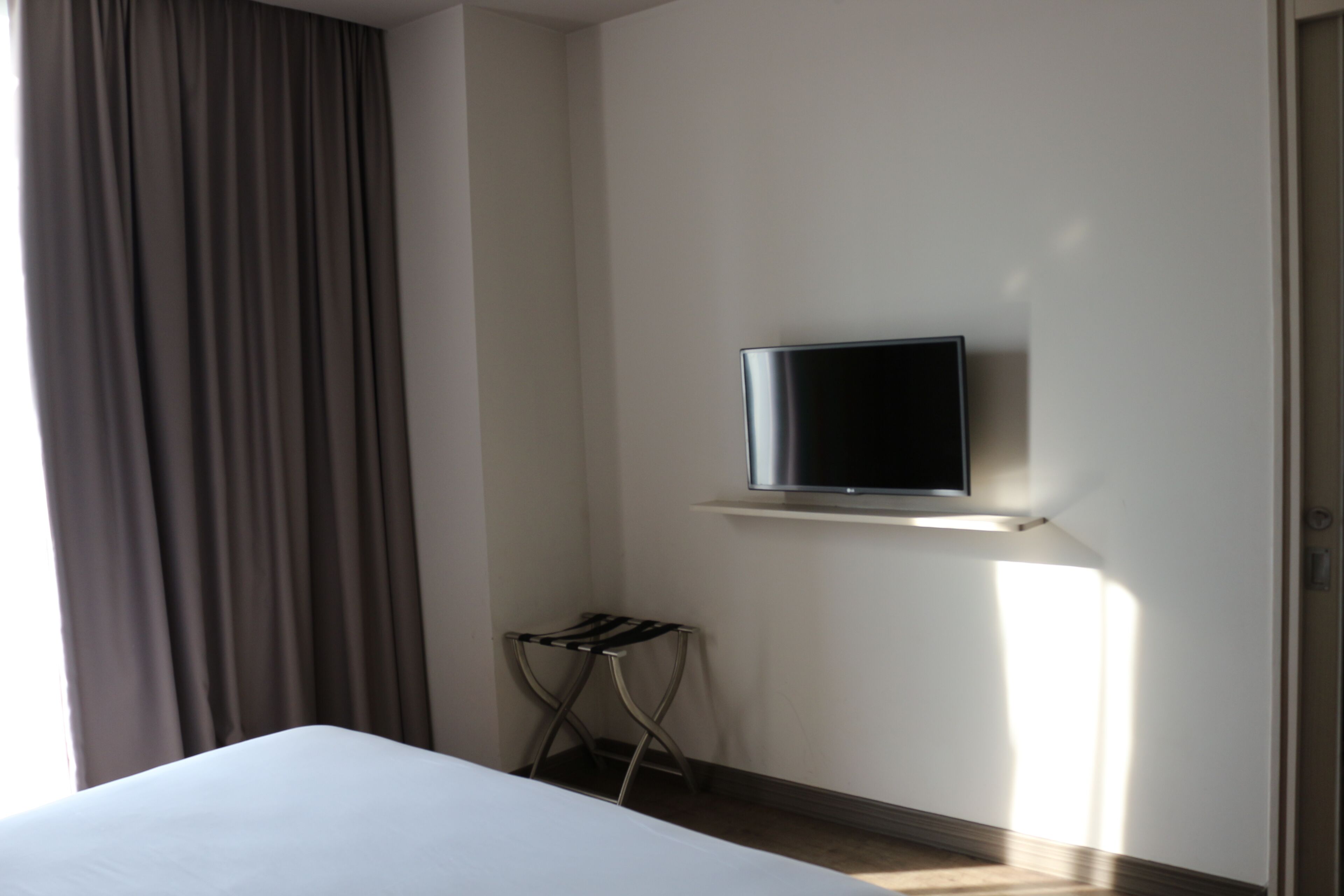 Hotellihuone, jossa on sänky, seinälle asennettu televisio, pieni pöytä ja ikkuna, jossa on verhot.