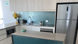 A modern kitchen with light wood cabinets, a stainless steel refrigerator, a built-in oven, and a teal island with a sink. - Nova Mooloolaba Beach (Mooloolaba)